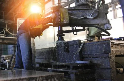 Craftsman uses a large machine to shape stone in a factory, illuminated by na Stock Photos