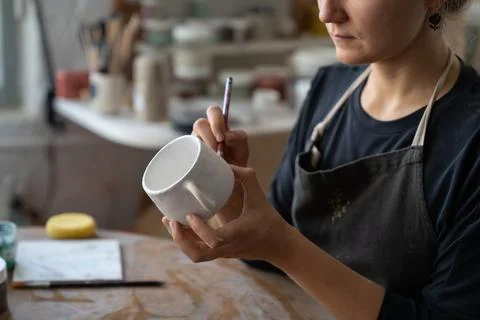 Craftswoman makes sketch of future pattern on white mug to decorate kitchen with Stock Photos