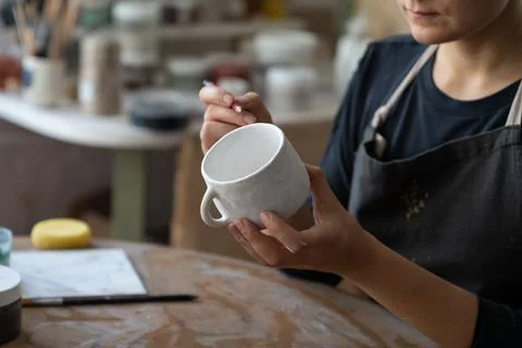 Craftswoman makes sketch of future pattern on white mug to decorate kitchen with Stock Photos