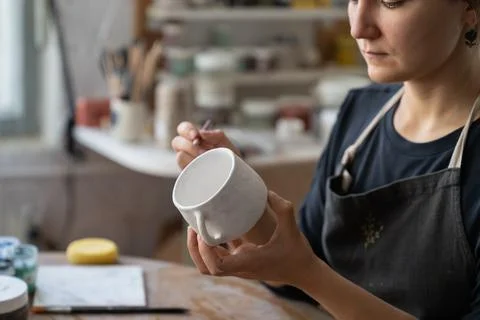 Craftswoman makes sketch of future pattern on white mug to decorate kitchen with Stock Photos