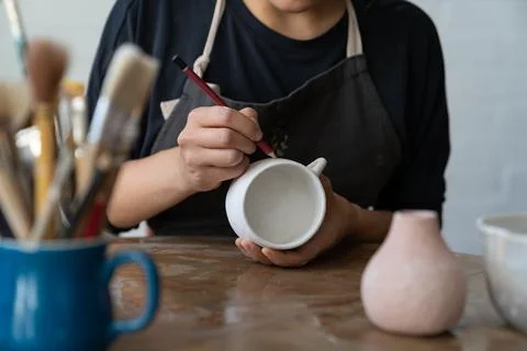 Craftswoman makes sketch of future pattern on white mug to decorate kitchen with Stock Photos