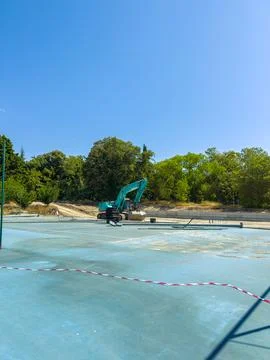 A crane is working on a construction site with a blue surface. Trees are visi Stock Photos