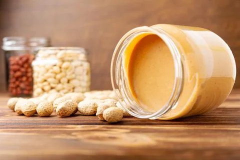 Creamy peanut paste in open glass jar, peanut butter spoon, peanuts and peanu Stock Photos