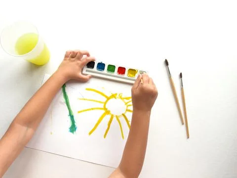 Creative child using watercolors to paint a bright sun with brushes and paper Stock Photos