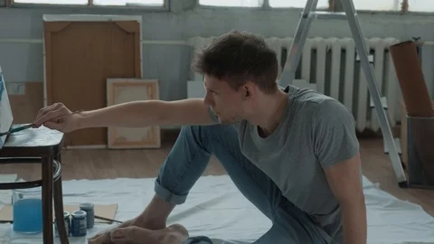 Creative man in bright workshop sits on the floor opposite a canvas Video stock 125613404
