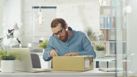 Creative Man Opens Up Parcel While Sitting in His Bright and Modern Office. Stock Footage 70627882