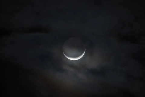 Crescent moon partly hidden by thin clouds in a dark night sky Stock Photos