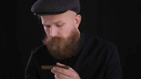 Criminal man in cap sniffs cigar on black background.close up,caucasian Stock Footage 130228077