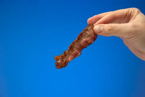 Crispy fried bacon strip or slice held between finger by Caucasian male hand. Fotos de archivo