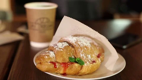 Croissant in a paper wrapper on a table. In the background is a glass of coffee. Stock Footage 83561551