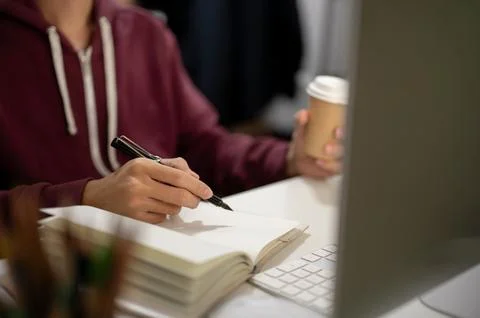 Cropped image of a man sitting in front of a computer, taking notes in a bo.. Foto stock