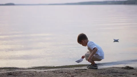 Crouching boy placing paper boat on calm lake water. Side view of child Stock Footage 336382407