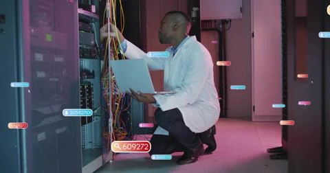Crouching IT tech in white coat checking colorful cables in server aisle, laptop Foto stock