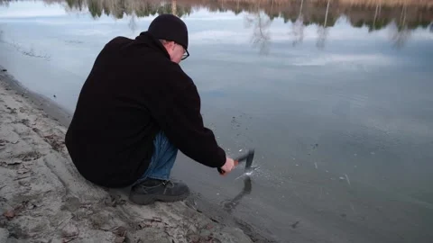 Crouching man breaking thin layer ice on lake with small axe. Stock Footage 321231543
