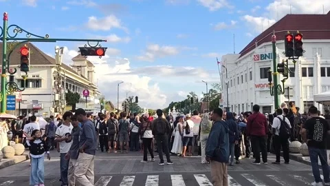 A crowd gathers at a crosswalk under sunny sky. Many stand, some sit, all w.. Stock-Footage 341997673