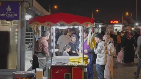 Crowds pass a red awning corn cart on a busy Istanbul sidewalk at night Vídeos de archivo 347793998