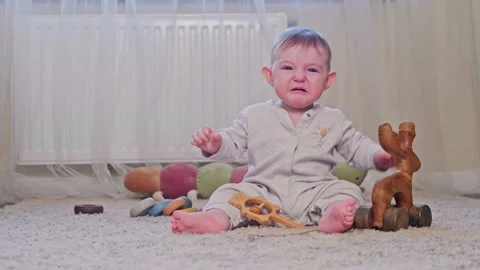 Crying baby boy sitting on light floor with wooden toy and rattle. Sad infant Stock Footage 337427313