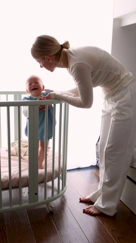 Crying baby standing in crib while mother gently comforts her. Real parenting Stock Footage 310046590