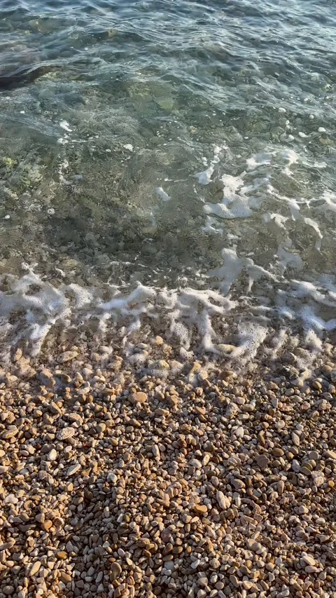 Crystal clear sea wave with soft foam gently washing over a sunny pebble beach Stock Footage 314947945