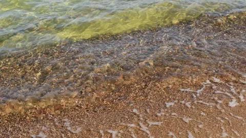 Crystal clear wave gently washes over pebbled shoreline under sunlight, creating Stock Footage 310168873