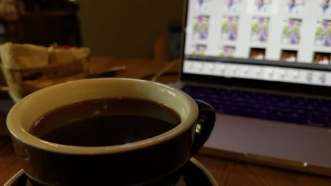 Cup of coffee on desk beside blurred laptop screen in warm home workspace light Stock Footage 345605562
