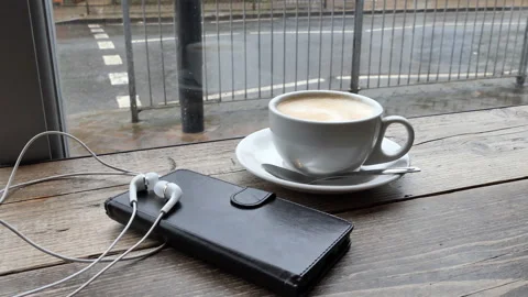 A cup of coffee, a mobile phone and headphones on a table. Raining outside. Stock Footage 89452853
