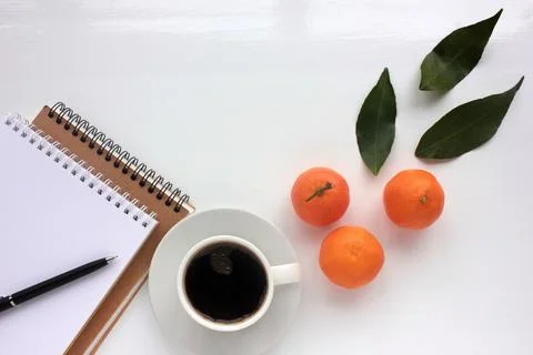 Cup of coffee, notebook, and oranges fruits with leaves on white office desk. Stock Photos