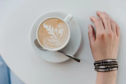 Cup of coffee with a pattern and a female hand with beads on a white table Stock Photos