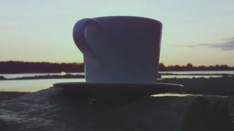 A cup of delicious hot tea or coffee on a large stone, with a steam at sunset by Stock Footage 80859446