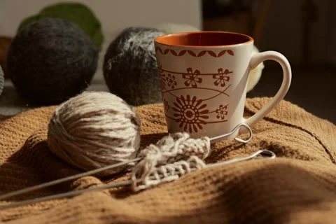 A cup sits beside soft yarn and knitting needles on a blanket. Sunlight shines Видео 336464813