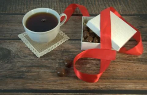 A cup of tea, a white box of chocolates with a red satin ribbon on a wooden t Stock Photos