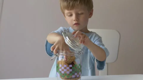 Curious 4-year-old blond boy is lifting the lid of a glass jar filled with Stock Footage 318293570
