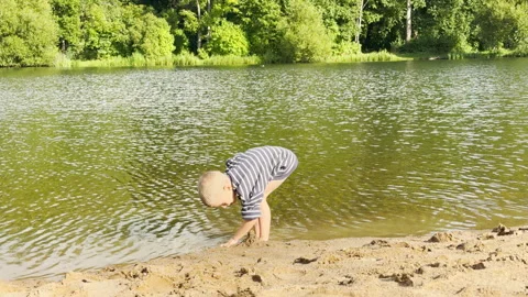 A curious boy bends over to play with sand at the water's edge. Video stock 344989672