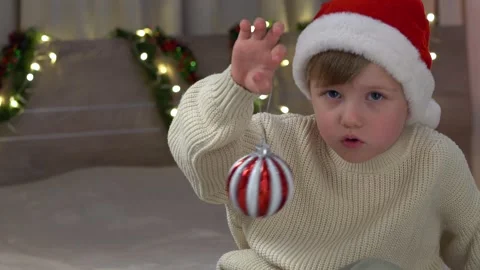 Curious boy in Santa hat examining red and white holiday ornament, interacting Stock Footage 322195699