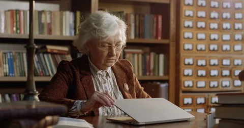 Curious Caucasian old gray-haired woman sitting at table in library and opening Video stock 125332705