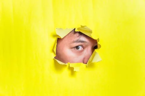A curious eye peeks through a torn yellow paper backdrop. Stock Photos