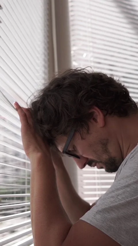 Curious man looking through window blinds and lowering his head in despair or Vídeos de archivo 348286425