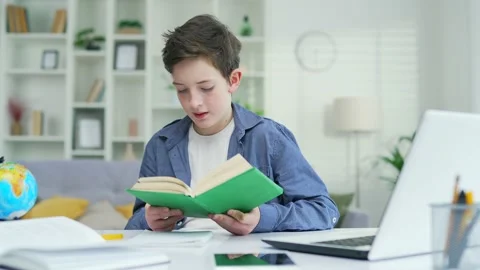 Curious school boy studies sitting at a table reading a book in a home classroom Stock Footage 238632210