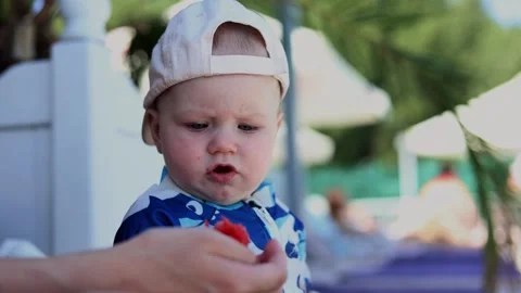 Curious toddler examines a small red snack offered by an adult outdoors in Stock Footage 347686484