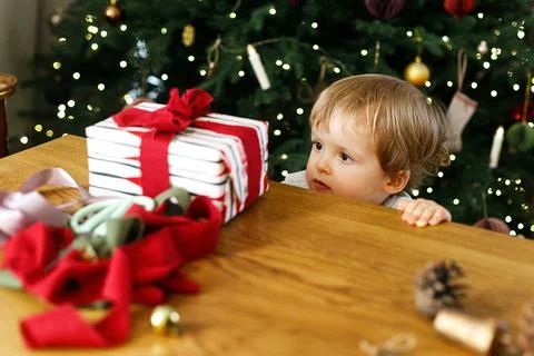 Curious Toddler Peeking over Table at Wrapped Christmas Gift near Glowing Tre 库存照片