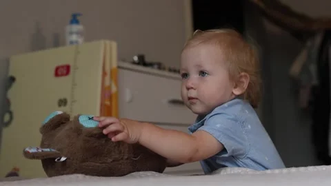 A curious toddler playing with a soft brown teddy bear on a bed in a cozy home Stock Footage 345610908