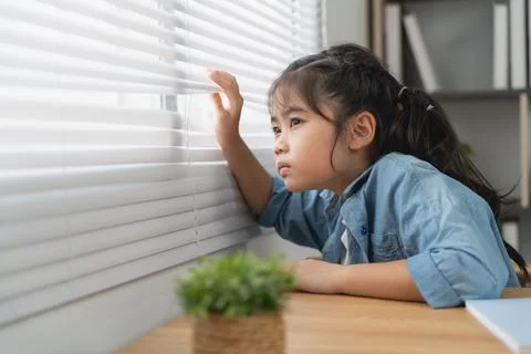 Curious young girl peeks through window blind in contemplative moment indoo.. Stock Photos