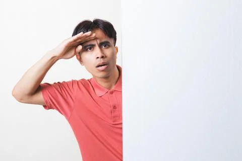 Curious young man peeking from behind a blank wall while shading his eyes a.. Stock Photos
