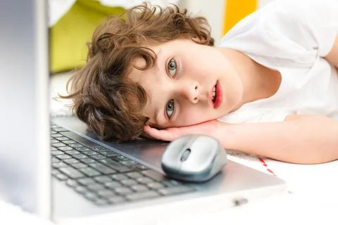 Curly boy is engaged at computer. Tired child lies covering his face with his Stock Photos