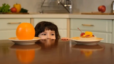 Curly boy peeking from the table and choosing between cake or an orange Stock Footage 125705828