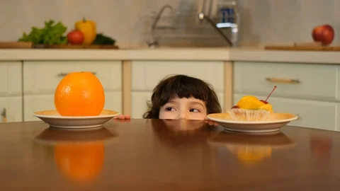 Curly boy peeking from the table and choosing cake or an orange Stock Footage 125706027