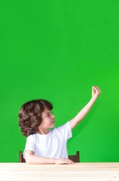 Curly cute boy sitting at a table half-turned his hand raised. Close-up. Gree Stock Photos