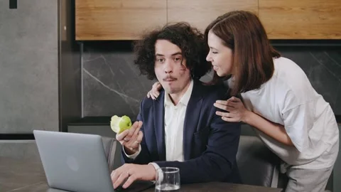A curly-haired businessman working remotely on his laptop at home, eating an Stock Footage 296832860