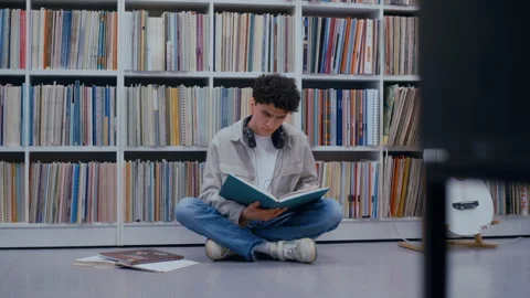 Curly-Haired College Guy on Library Floor Reading Book While Wearing Headphones Stock Footage 335124084
