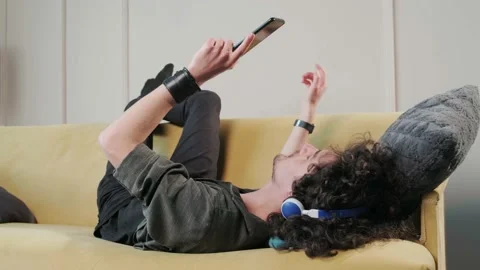Curly haired guy on the couch listening music with headphones, having fun. Stock Footage 155807201